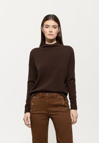 Frau mit glattem, braunem Haar, die einen dunkelbraunen Rollkragenpullover und hochgeschnittene braune Hosen mit goldenen Knöpfen trägt.