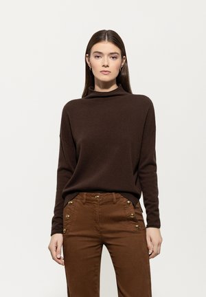 Woman with straight brown hair wearing a dark brown turtleneck sweater and high-waisted brown pants with gold buttons.