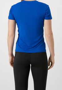 Person står vänd bort med en åtsittande blå kortärmad skjorta och svarta byxor mot en enkel bakgrund.