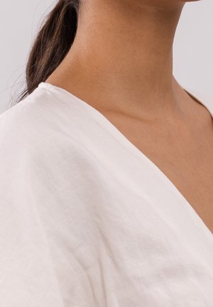 Gros plan sur le cou et l'épaule d'une personne portant un vêtement blanc à col en V, avec des cheveux foncés attachés en arrière.