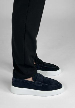 Navyblauwe suède loafers met een witte plateauzool, voorzien van een gestikt ontwerp en een banddetail aan de voorkant, gedragen met zwarte broeken.