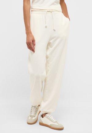 Person trägt cremefarbene Jogginghose mit Kordelzug in der Taille und elastischen Bündchen, kombiniert mit weißen und beigen Sneakern.