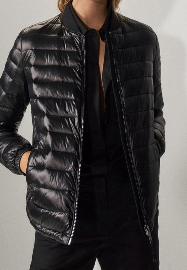 Personne portant une veste bomber matelassée noire brillante sur une chemise noire partiellement déboutonnée, mains dans les poches de la veste, fond neutre.
