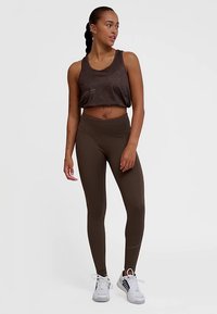 Brązowy krótki top z geometrycznym wzorem, zestawiony z brązowymi legginsami z wysokim stanem. Modelka nosi białe trampki, prezentując dopasowany sportowy wygląd.
