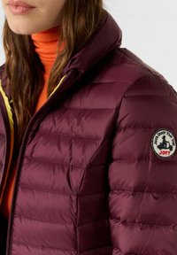 Chaqueta acolchada burdeos con cuello alto, con interior amarillo y un parche redondo en la manga izquierda.