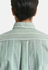 Camicia verde a righe con colletto a punta, dettaglio con piega posteriore e bottone al colletto. Il tessuto sembra leggero e ha una trama liscia.