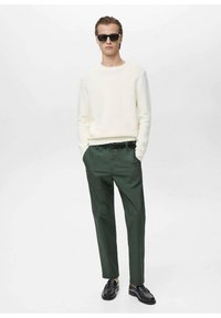 White ribbed sweater, green trousers, black belt, and black loafers. Model wears black sunglasses. Minimalist design, fitted silhouette.