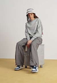 Felpa grigia con logo, pantaloni a gamba larga grigi e sneakers blu/bianche. Cappello a secchiello a righe. Seduto su una valigia metallica con rotelle.