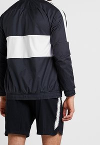 Zwart-witte windbreaker jas met een kleurblokontwerp, featuring een grote witte streep over de rug. Gemaakt in combinatie met zwarte shorts.