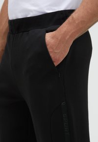 Schwarze Sporthose aus glattem Stoff mit einem bequemen Bund, einer Reißverschlusstasche und einem minimalistischen "SPORT"-Markierungsdetail.