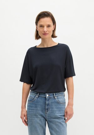 Femme aux cheveux bruns courts se tenant de face, portant un haut noir ample à manches courtes et un jean bleu sur un fond blanc uni.