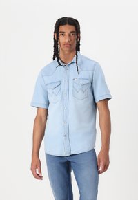 Lys blå denim kortermet skjorte med to brystlommer med spissere klapper, trykknapper og kontrasterende sømdetaljer.