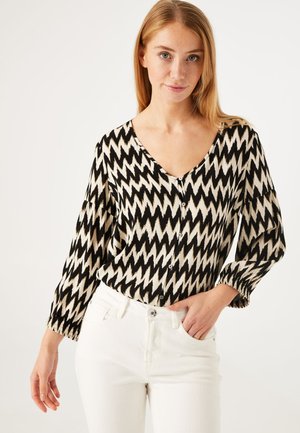 Zwarte en crèmekleurige blouse met patroon, V-hals, lange mouwen, knopen over de voorkant en elastische manchet, gecombineerd met witte jeans.