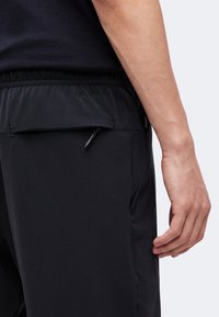 Nahaufnahme einer Person, die schwarze Dri-Fit-Sporthose mit Reißverschlusstasche hinten und elastischem Bund trägt, Hand ruht an der Seite.