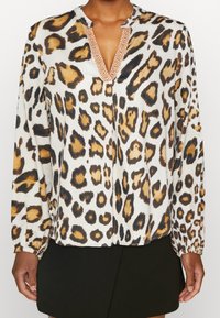 Blusa a maniche lunghe con stampa leopardata in bianco, marrone e nero. Presenta una scollatura a V con dettagli ricamati e polsini elasticizzati.