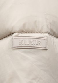 Pufinė striukė šviesiai smėlinės spalvos audinio, su išsikišusiu baltu guminiu logotipo ženkleliu, pažymėtu "HOLLISTER", ant glotnaus paviršiaus.