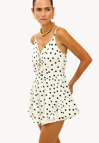 Robe blanche à pois noirs, avec un décolleté en V, des bretelles réglables et un ourlet volanté. Le tissu semble léger et fluide.