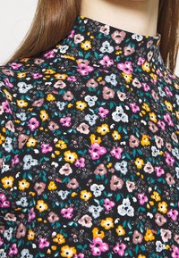 Nahaufnahme eines hochgeschlossenen schwarzen Stoffs mit kleinen rosa, gelben, weißen und braunen Blumenmustern sowie grünen Blättern.
