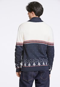 Stickad tröja med en krämfärgöverdel och marineblå underdel, med ett rödvitt zigzag-mönster och vita snöflinge- och trädmotiv vid nederkanten.