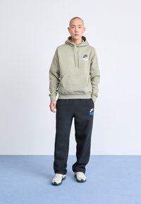 Olivno zeleno hoodie s kenguru žepom i belim logom, uparen s crnim trenirkama koje imaju logo i sivo/belim patikama.