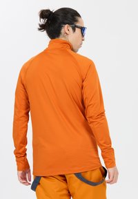 Langärmliges Shirt in leuchtendem Orange, mit hohem Kragen und glatter Textur. Die Rückseite zeigt Raglanärmel und nahtlose Designdetails.