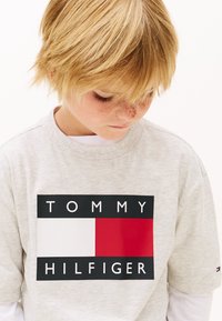 Grå bomullst-shirt med en framträdande logotyp med röda, vita och blå rektanglar samt texten "TOMMY HILFIGER" i svart.