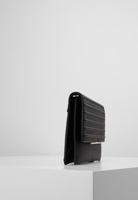 Svart läderclutch med quiltat lock och guldmetalldetalj, stående upprätt på en vit yta mot en enkel grå bakgrund.