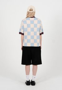 Person iført en blå og beige ternet kortærmet skjorte, sorte shorts, hvide sokker, sorte sko og en lys beige kasket, der står med ryggen til.