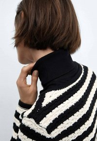 Pull rayé en fil noir et blanc ; il présente un design tricoté texturé et un col haut à côtes. Une main touche le col par derrière.