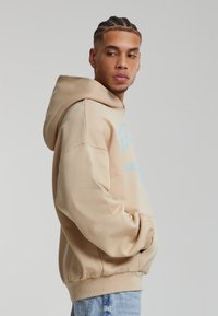 Felpa oversize beige con tasca a canguro, caratterizzata da scritte stampate azzurre e polsini a coste. Tessuto morbido con vestibilità comoda.