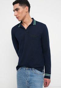 Navy long-sleeve polo shirt with a green and white striped collar, featuring embroidered logo and button closure, paired with light blue jeans.
