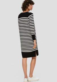 Schwarz-weiß gestreiftes Strickkleid mit langen Ärmeln, seitlichem Schlitz und schwarzem Saum. Kombiniert mit weißen Sneakern.