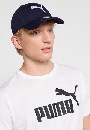 Joven con una gorra azul marino de Puma y una camiseta blanca de Puma sobre un fondo claro y liso, mirando ligeramente hacia un lado.