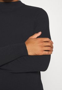 Svart, ribbad polotröja som visas med korsade armar, vilket framhäver åtsittande ärmar och mjuk textur.