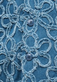 Ύφασμα τζιν με υφή κεντήματος σε βρόχους και δύο σκούρα μπλε κουμπιά με τέσσερις τρύπες ραμμένα σε κάθετη σειρά.