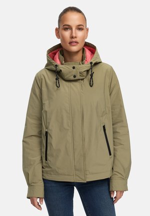 Vrouw draagt een olijfgroene jas met capuchon en koraalkleurige voering, zwarte zakken met rits, en blauwe spijkerbroek, staande tegen een effen achtergrond.