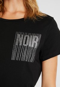 Black short-sleeve t-shirt featuring a silver metallic "NOIR" text design with vertical stripes, showcasing a smooth fabric texture.