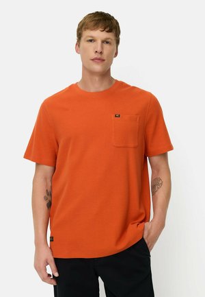Mand med kort hår iført en orange kortærmet skjorte med brystlomme og sorte bukser, stående med den ene hånd i lommen.