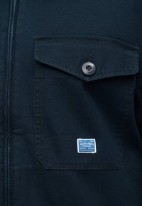 Σκούρο μπλε ύφασμα με τσεπάκι με κλείσιμο με κουμπιά και μια μικρή μπλε και λευκή ετικέτα "Jack & Jones Premium" κάτω από το τσεπάκι σε ένδυμα.