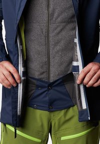 Giacca da marinaio con foderatura in pile grigio, dotata di zip frontale e chiusura a scatto. I pantaloni verdi hanno tasche con zip brillanti e una texture resistente.