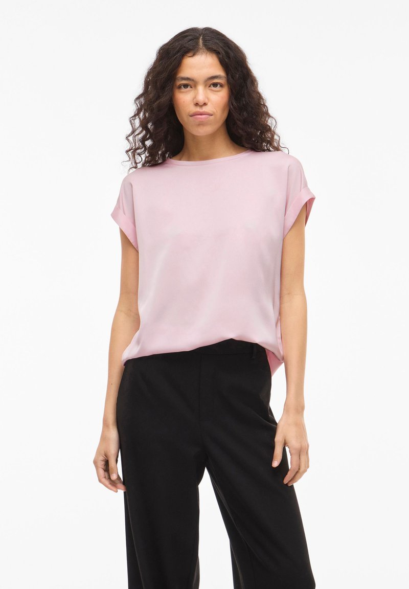 Femme aux cheveux bouclés portant un chemisier rose clair à manches courtes et un pantalon noir, debout devant un fond blanc.