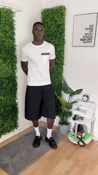 Young man stands with hands behind back between two vertical green plant panels, next to a plant, sewing machine, and framed motivational quote.
