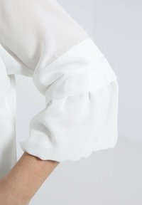 Weiße Bluse mit Ärmeln aus einem weichen, durchsichtigen Stoff. Puffdetail am Bündchen sorgt für eine lockere Passform. Glatte Textur mit einem klaren, minimalen Design.