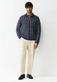 Tmavomodrý pletený kabát s mriežkovým vzorom, so zapínaním na zips a golierom. Kombinovaný s krémovými nohavicami a čiernymi textúrovanými topánkami.