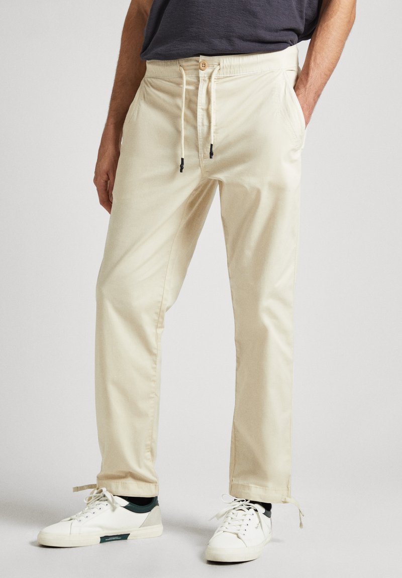 Pantalones de mezcla de algodón beige con cintura elástica y cordones, que presentan bolsillos laterales y piernas ajustadas; combinados con zapatillas blancas.