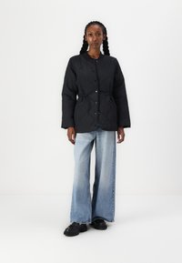 Veste noire matelassée avec une taille cintrée et une fermeture à boutons. Assortie avec un jean taille haute bleu clair à pattes d'élargissement et des chaussures noires. Texture lisse sur l'ensemble.