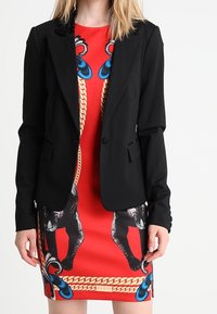 Veste noire ajustée, coupe cintrée, fermeture à un bouton, associée à une robe rouge ornée d'imprimés animaliers et de motifs de chaînes bleues. Texture lisse.