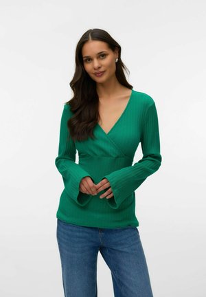 Pikkade tumedate juustega naine, kes kannab rohelist soonikutega V-kaelusega mähistoppi, millel on lehvivate varrukatega, ja siniseid teksapükse, seisab valge tausta ees.