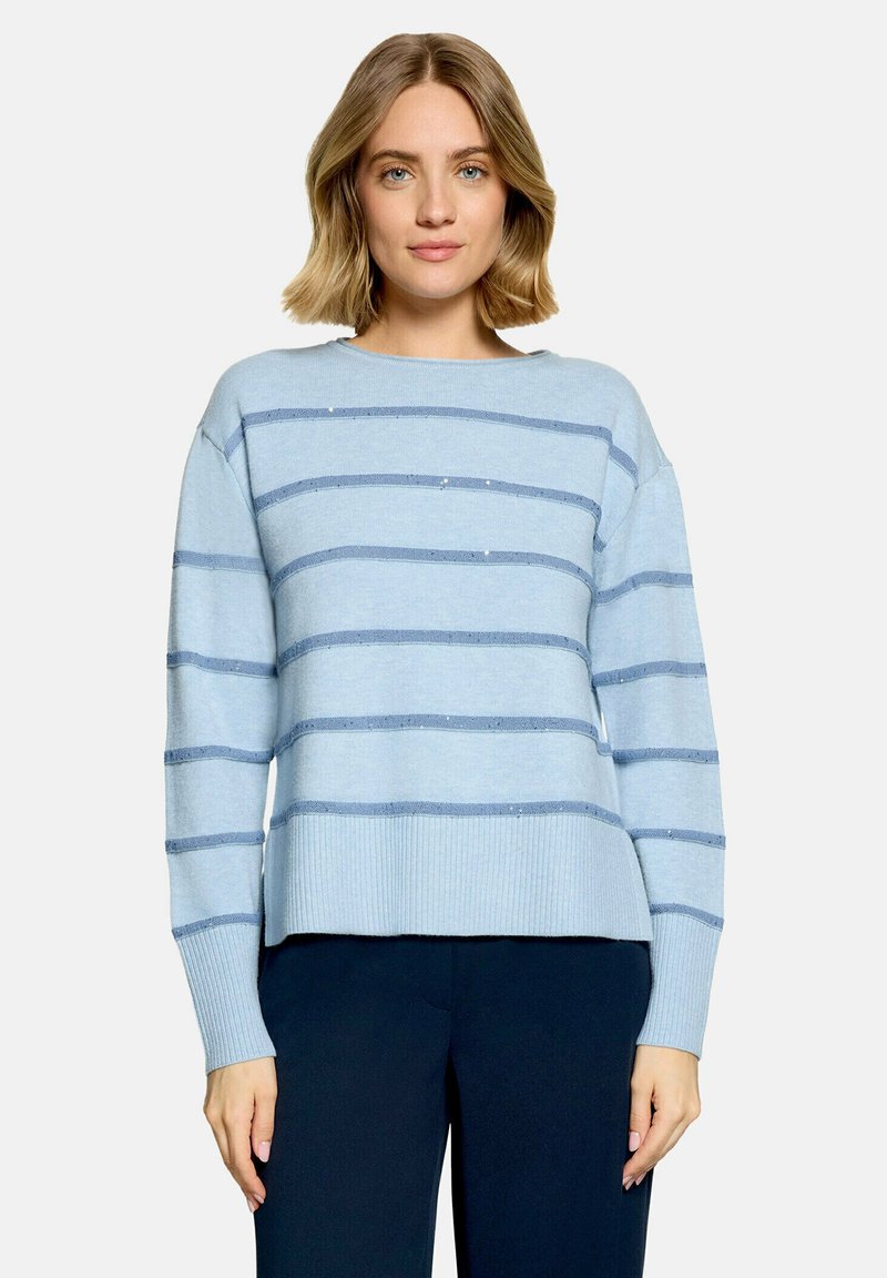 Femme portant un pull bleu clair avec des rayures horizontales bleu foncé et un pantalon bleu foncé, debout devant un fond clair uni.