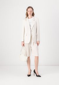 Blazer créme sur t-shirt graphique blanc, associé à un short beige clair. Des escarpins noirs à talons pointus et un sac à bandoulière blanc complètent le look.